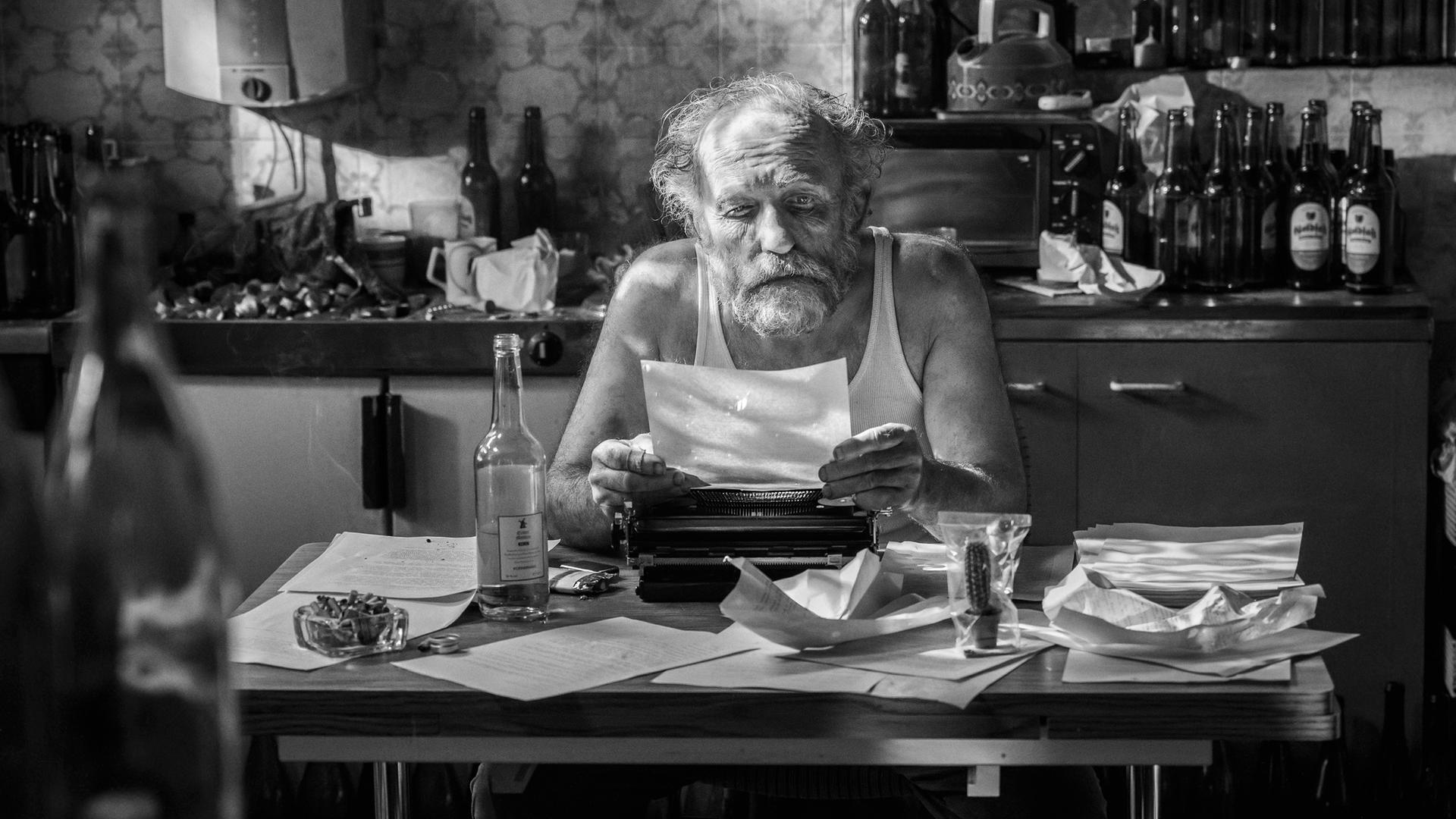 Man sieht die schwarz-weiß-Fotografie eines älteren Mannes, der an einem Tisch mit einer Schreibmaschine sitzt, ein Blatt Papier in den Händen hält und liest. Auf dem Tisch liegen viele Blätter neben einer Flasche, einem Aschenbecher und einem kleinen Kaktus in einer Plastiktüte.