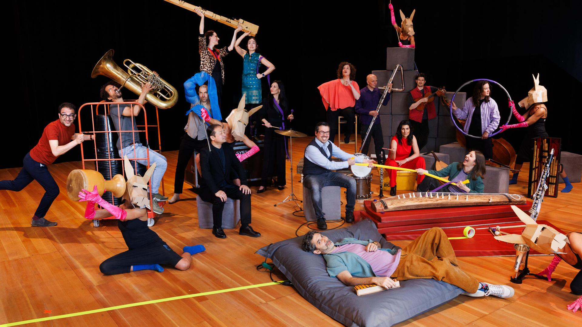 Man sieht Musikerinnen und Musiker mit ihren Instrumenten auf einer Bühne. Manche tragen Tierkopfmasken, spielen Instrumente und benutzen diverse bunte Alltagsgegenstände.