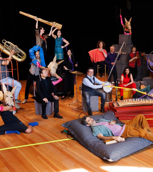 Man sieht Musikerinnen und Musiker mit ihren Instrumenten auf einer Bühne. Manche tragen Tierkopfmasken, spielen Instrumente und benutzen diverse bunte Alltagsgegenstände.