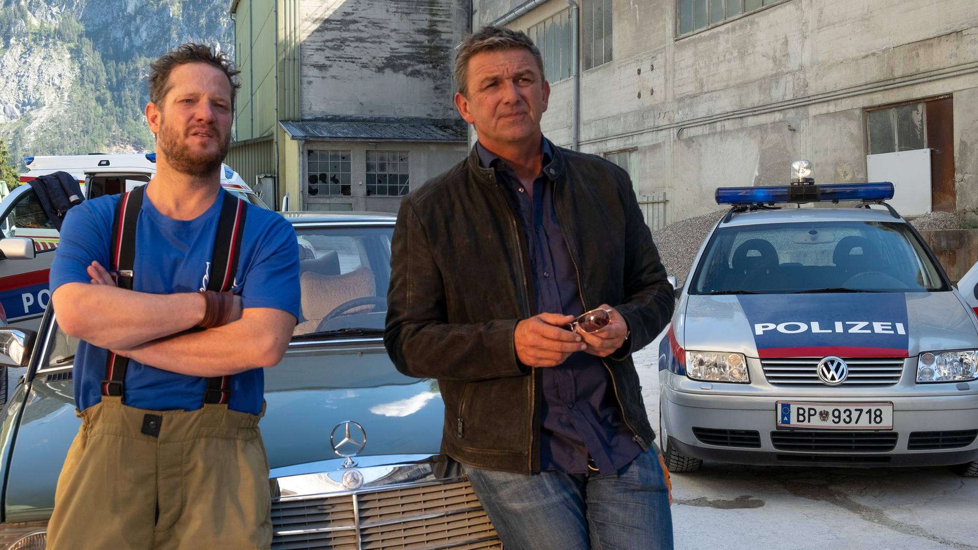 Martin (Hans Sigl, r.) versucht, Andreas Kloser (Aurel Manthei, l.) ins Gewissen zu reden, der sich einfach nicht schonen will. Die beiden lehnen an der Motorhaube eines Autos und schauen in die Ferne.