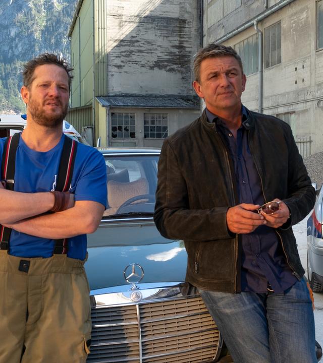Martin (Hans Sigl, r.) versucht, Andreas Kloser (Aurel Manthei, l.) ins Gewissen zu reden, der sich einfach nicht schonen will. Die beiden lehnen an der Motorhaube eines Autos und schauen in die Ferne.