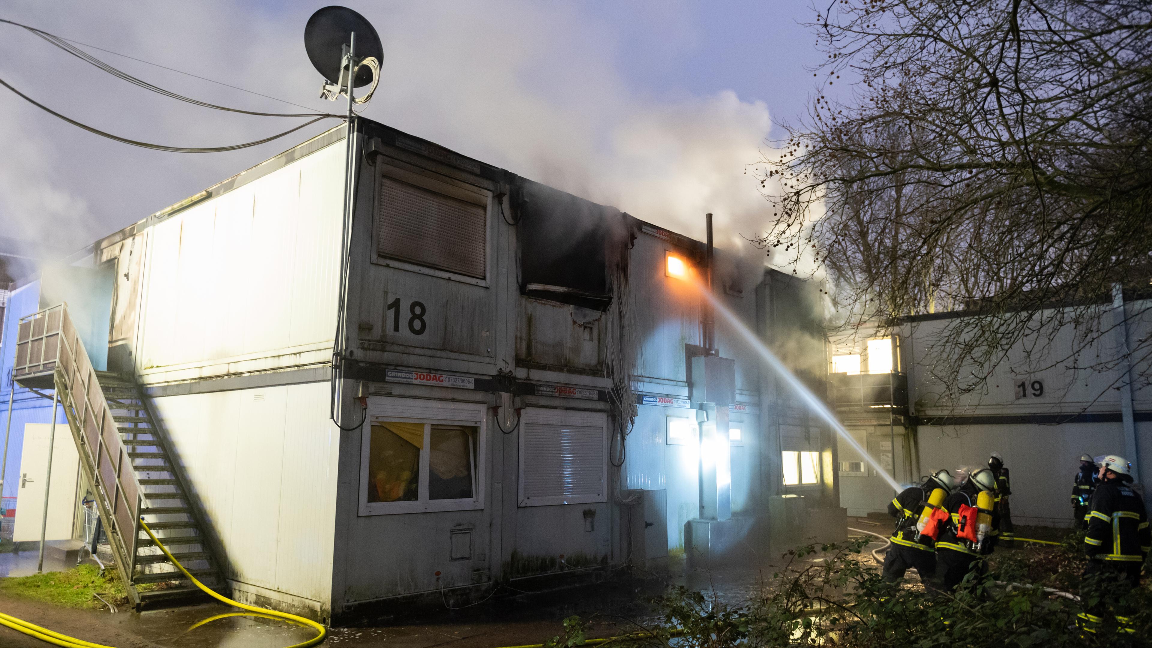 Hamburg: Feuerwehrleute stehen bei Löscharbeiten vor den verqualmten und teils brennenden Container-Einheiten einer Flüchtlingsunterkunft in Hamburg-Winterhude.