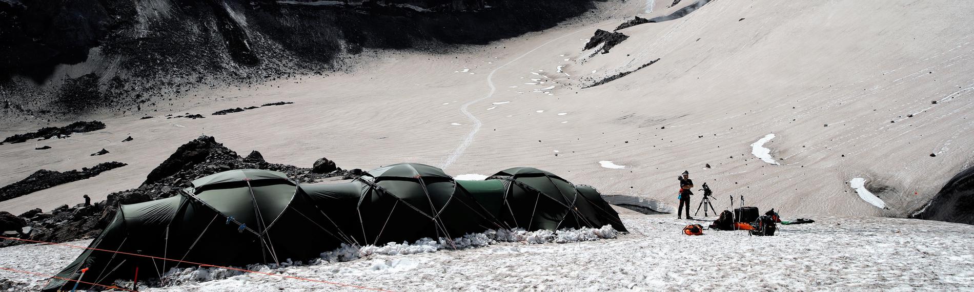 Großes, dunkles Expeditionszelt mit Männern davor, aufgebaut in einer Vulkanlandschaft. Das Gelände steigt zum Kraterrand an, blauer Himmel mit Wolken