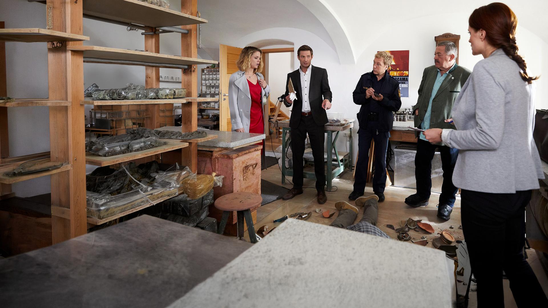 "Die Rosenheim-Cops - Feuer und Flamme": Anne Birkmeier (Sigrid Spörk) steht mit den Ermittlern Hansen (Igor Jeftic), Mohr (Max Müller), Hofer (Joseph Hannesschläger) und Mai (Sina Wilke) im Halbkreis um das Opfer herum, das am Boden liegt. Auf dem Boden verteilt liegen zahlreiche Keramikscherben.