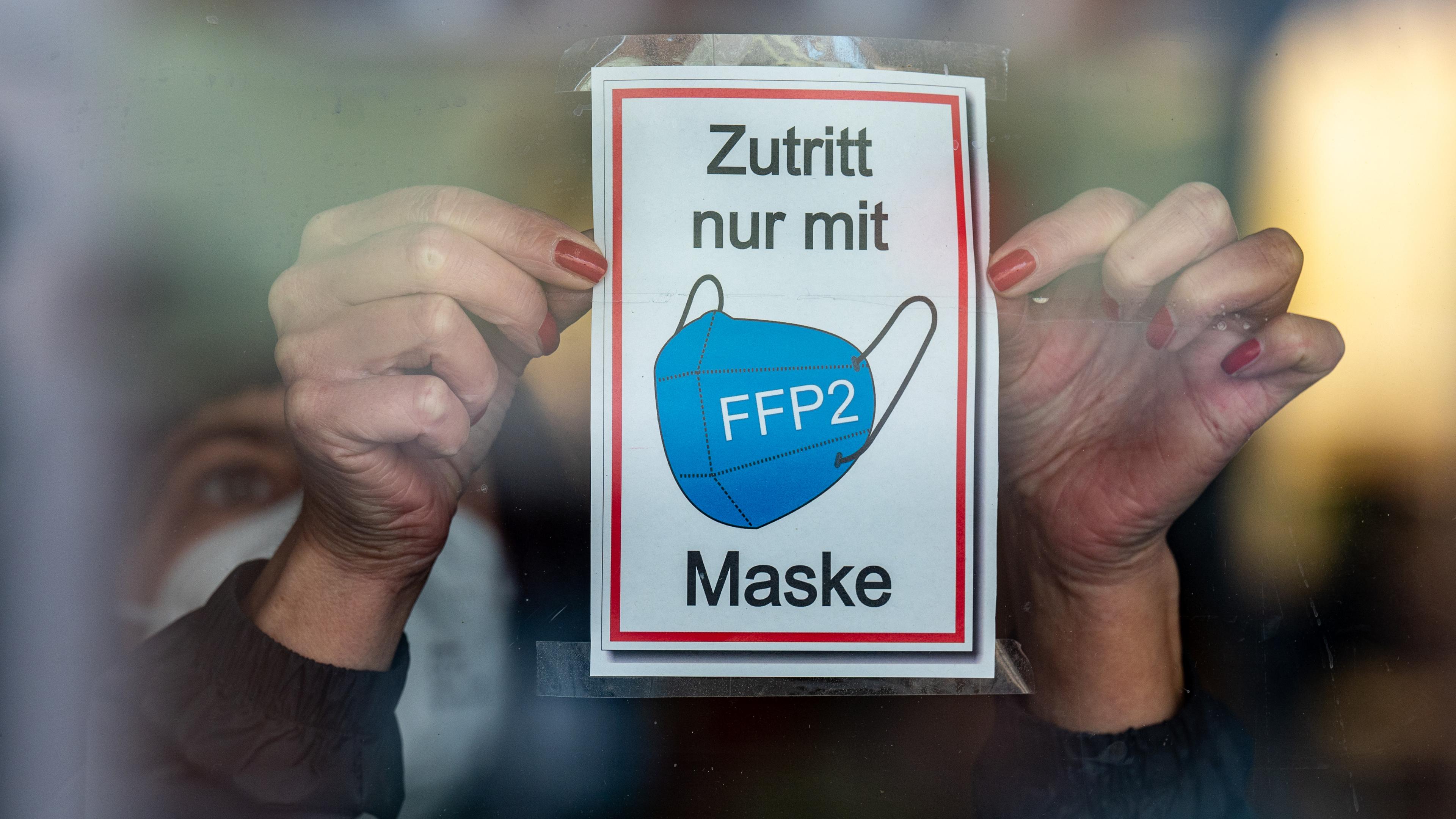  Bayern, Straubing: Eine Frau klebt ein Hinweisschild mit der Aufschrift ·Zutritt nur mit FFP2 Maske· an eine Glastür am Eingang eines Geschäfts in der Innenstadt.