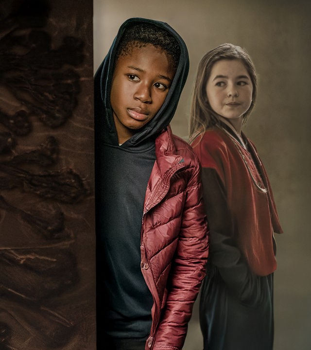 Nick (Aaron Sansi), ein etwa zwölfjähriger, schwarzer Junge, schaut im Flur eines Wohnhauses mit ernstem Gesicht um eine Ecke. Die Kapuze seines Hoodies hat er über seinen Kopf gezogen. Seine neue Mitschülerin Leni (Amelie Klein) steht unbemerkt rechts hinter ihm und schaut in seine Richtung.