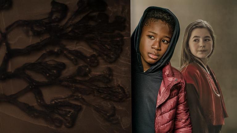 Nick (Aaron Sansi), ein etwa zwölfjähriger, schwarzer Junge, schaut im Flur eines Wohnhauses mit ernstem Gesicht um eine Ecke. Die Kapuze seines Hoodies hat er über seinen Kopf gezogen. Seine neue Mitschülerin Leni (Amelie Klein) steht unbemerkt rechts hinter ihm und schaut in seine Richtung.