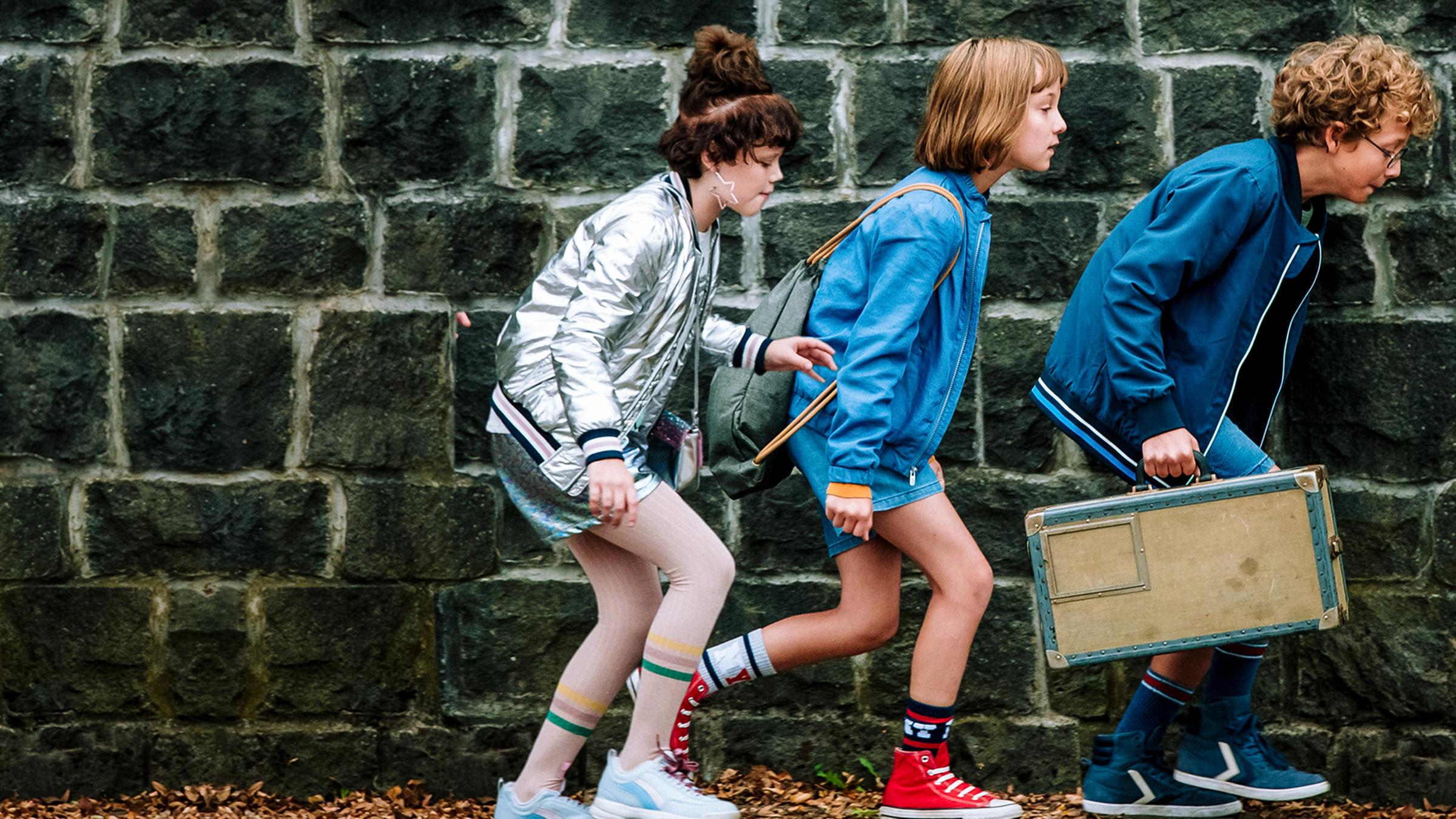 Cheyenne, Lotta und Paul schleichen in leicht gebückter Haltung an einer hohen Gartenmauer entlang. Paul trägt einen kleinen Koffer bei sich.