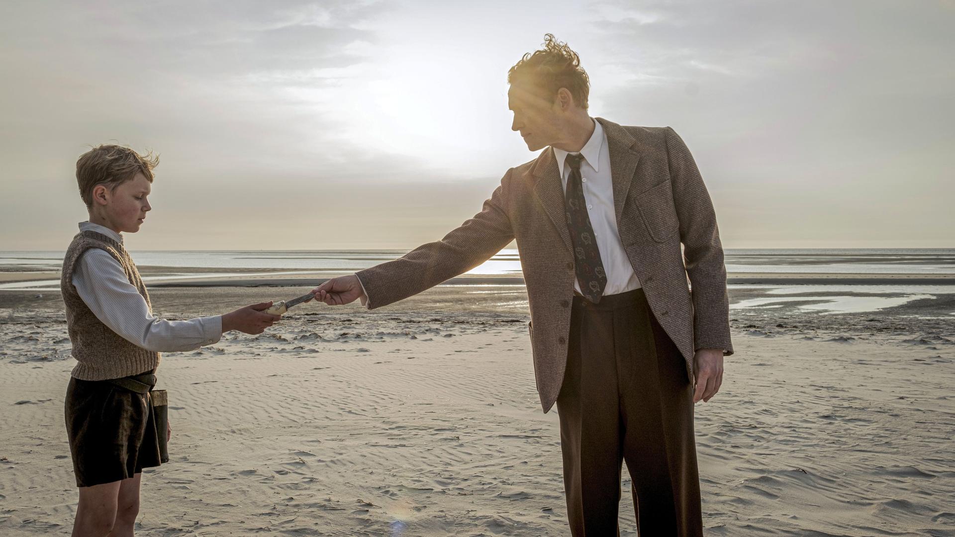 Filmszene: An einem Strand reicht ein Mann einem Jungen einen kleinen Gegenstand, der schlecht zu sehen ist. Beide tragen altmodische Kleidung aus der Nachkriegszeit. 
