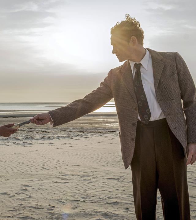 Filmszene: An einem Strand reicht ein Mann einem Jungen einen kleinen Gegenstand, der schlecht zu sehen ist. Beide tragen altmodische Kleidung aus der Nachkriegszeit. 