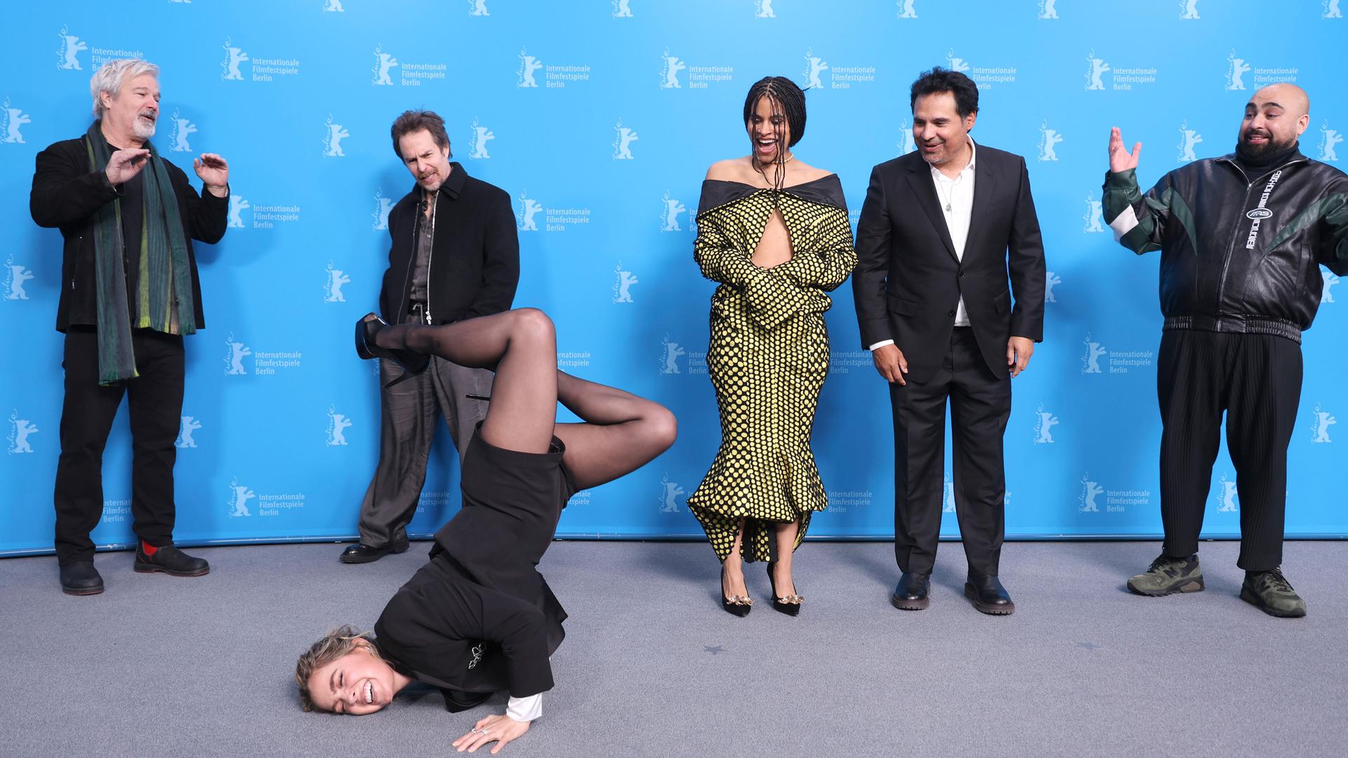 Vor dem blauen Hintergrund der Berlinale-Pressekonferenzen stehen mehrere Personen und schauen lachend zu einer Frau im schwarzen Anzug mit kurzen Hosen und Stöckelschuhen, die auf dem Boden Break-Dance vorführt.