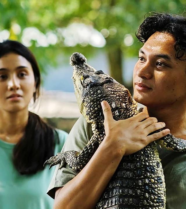 Ein junger  Mann trägt ein kleines Krokodil auf dem Arm, links hinter ihm steht eine junge Frau.