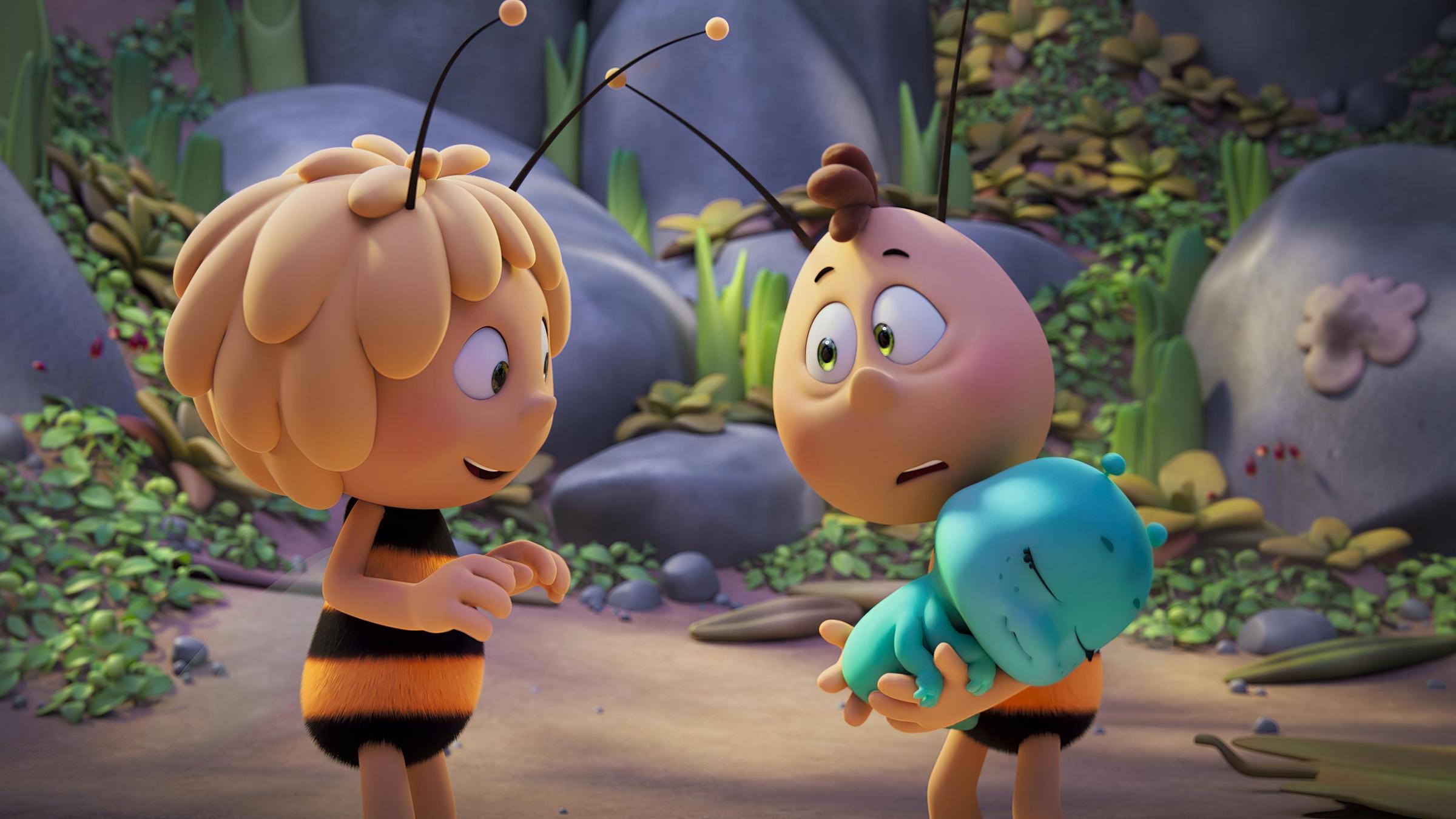 "Die Biene Maja - Das geheime Königreich": Zwischen große Felsbrocken wachsen grüne Pflanzen. Davor stehen sich Maja und Willie gegenüber. Maja trägt große Fühler auf dem Kopf, sie ist blond, hat ein kugelrundes Gesicht und einen schwarz-gelbgestreiften Pelz vom Hals bis zu den Beinen. Sie lacht Willie an. Der sieht wenig glücklich, ja eher hilflos aus, denn er trägt in seinen Armen die selig schlafende Ameisenprinzessin.