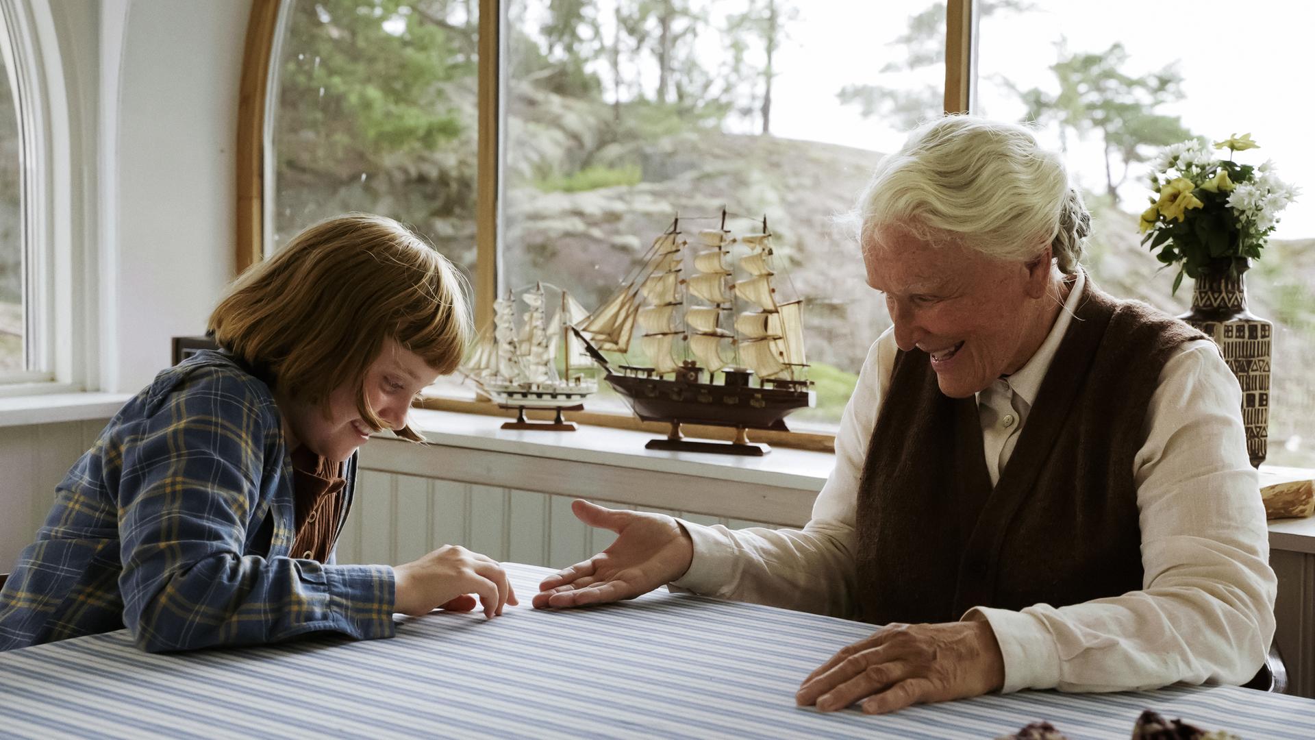 Ein junges Mädchen und eine alte Frau sitzen an einem Tisch. Beide lachen und blicken auf ihre Hände, die sie auf dem Tisch aufeinander zubewegen. Im Hintergrund ist ein Fenster zu sehen und auf dem Fensterbrett steht ein Modellschiff. 