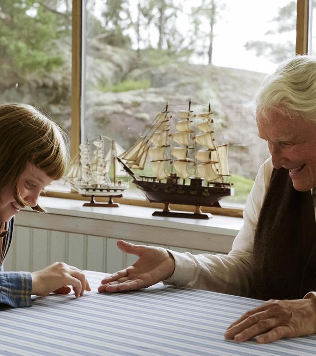 Ein junges Mädchen und eine alte Frau sitzen an einem Tisch. Beide lachen und blicken auf ihre Hände, die sie auf dem Tisch aufeinander zubewegen. Im Hintergrund ist ein Fenster zu sehen und auf dem Fensterbrett steht ein Modellschiff. 