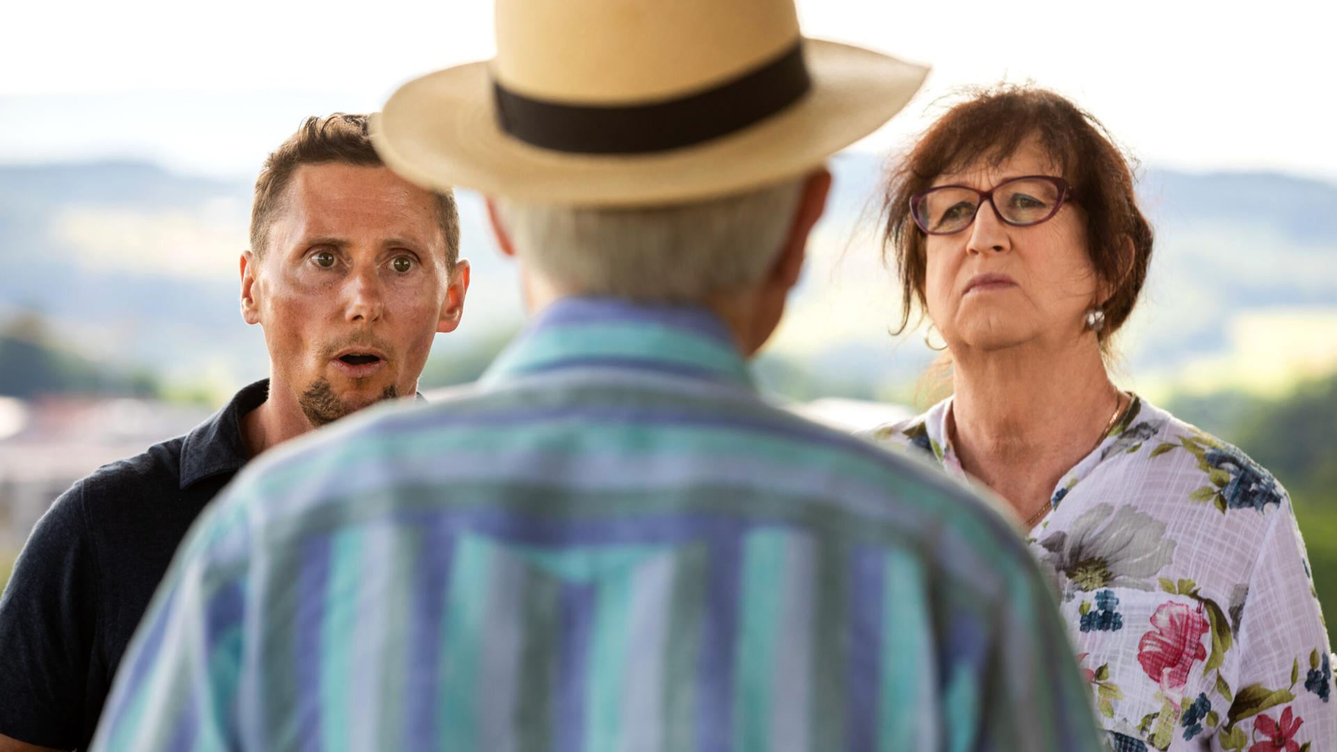 Man sieht einen Mann mit Strohhut von hinten. Links von ihm steht ein Mann, rechts von ihm eine Frau. 