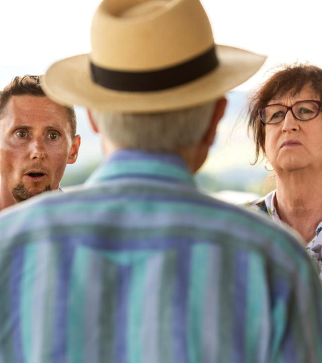 Man sieht einen Mann mit Strohhut von hinten. Links von ihm steht ein Mann, rechts von ihm eine Frau. 