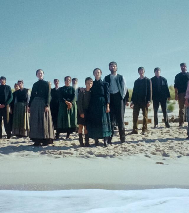 Eine Gruppe Menschen in historischer Bekleidung steht am Strand und schaut aufs Meer.