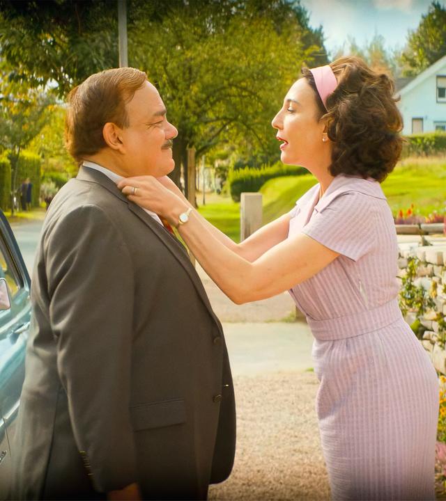 Ein Mann und eine Frau stehen einander gegenüber und sind für den Betrachter deswegen nur von der Seite zu sehen. Der Mann trägt einen Anzug und die Frau ein pastellfarbenes lila Kleid mit passendem Haarband. Die Frau richtet den Hemdkragen des Mannes, während sie mit ihm redet.