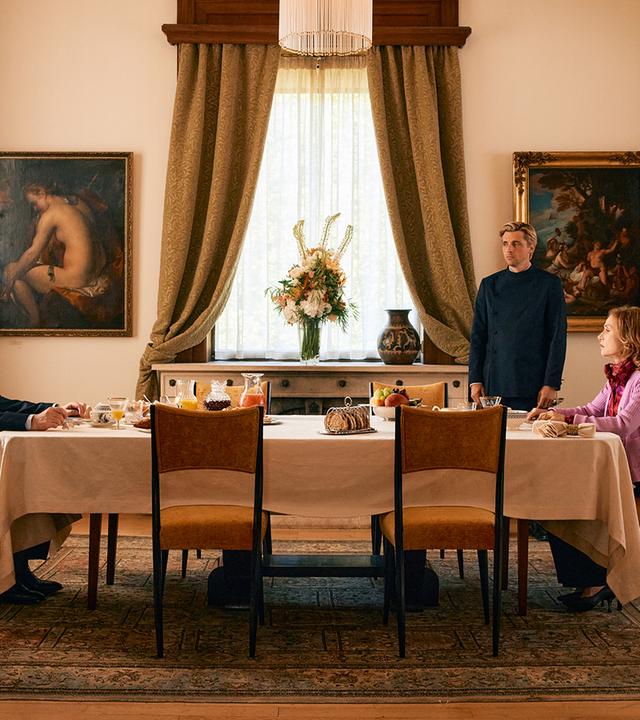 Man sieht eine Tafel an der ein Mann und eine Frau sitzen. Ein Butler steht neben dem Tisch.