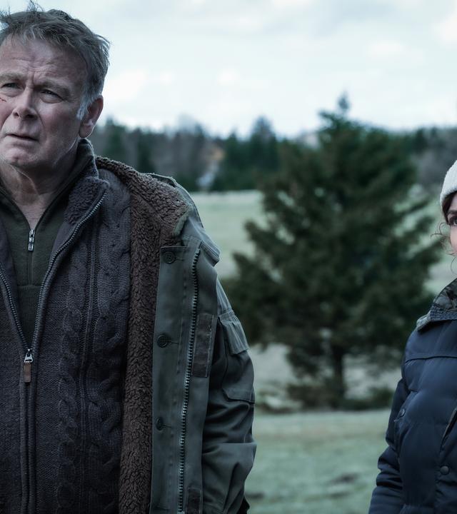 Ein Mann und eine Frau stehen in winterlicher Kleidung vor einem Waldstück.