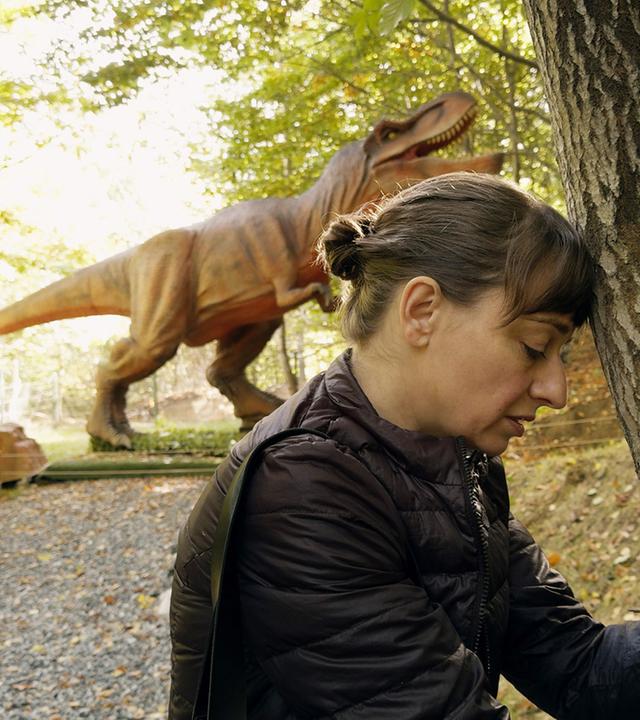 Eine Frau lehnt ihren Kopf an einen Baum und im Hintergrund steht ein Dinosaurier.