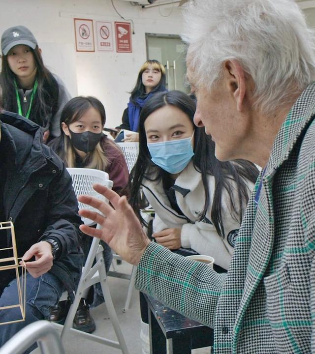 Man sieht einen älteren Herren, der mit einer Gruppe junger asiatisch aussehender Menschen spricht.