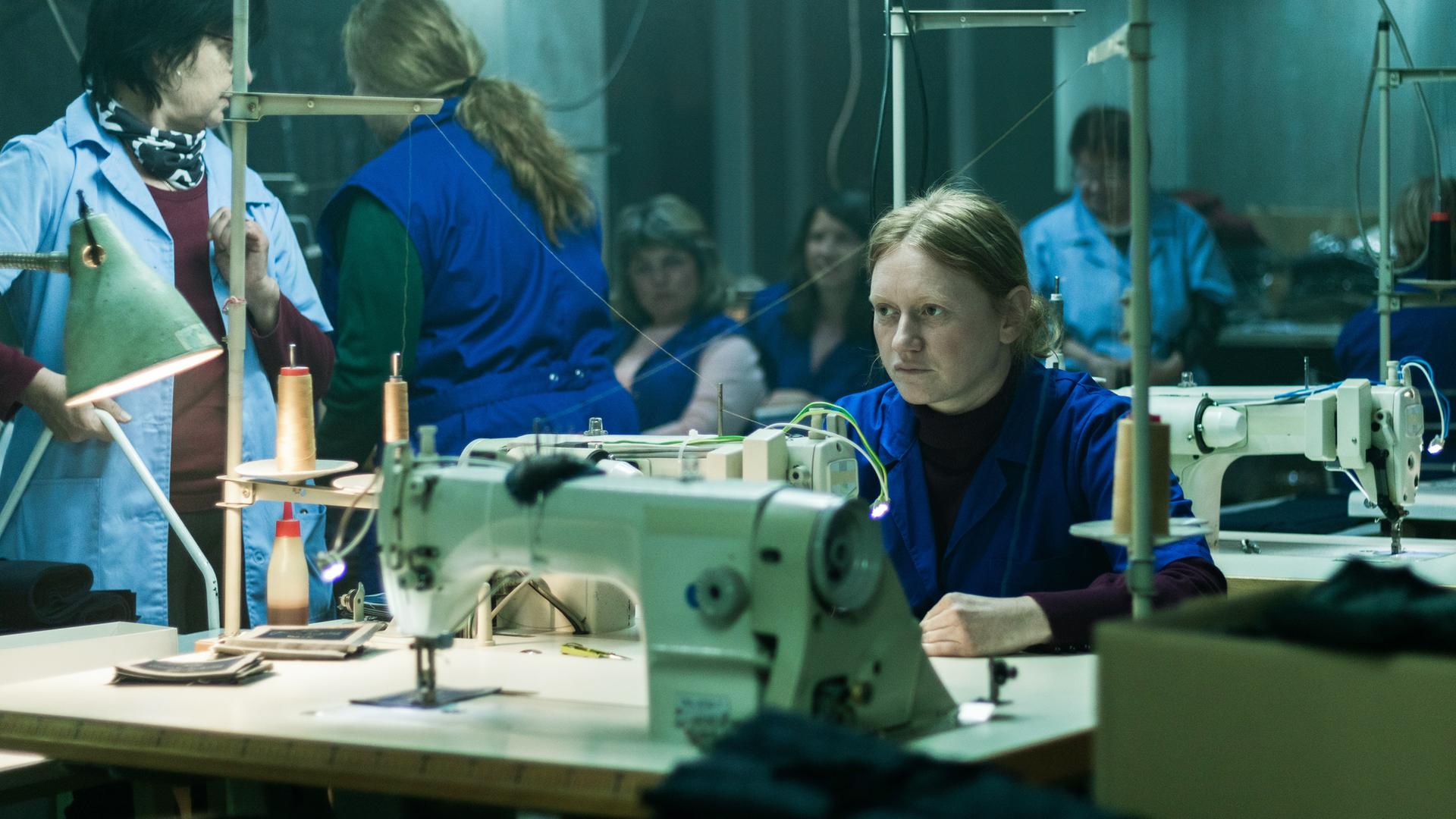 Eine blonde Frau in einem blauen Kittel sitzt in einer Werkshalle an einer Nähmaschine und schaut nachdenklich in die Ferne.