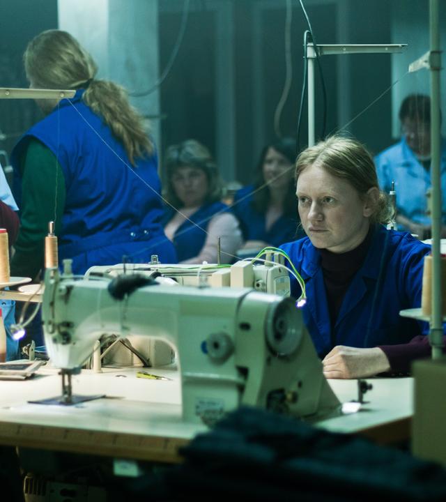 Eine blonde Frau in einem blauen Kittel sitzt in einer Werkshalle an einer Nähmaschine und schaut nachdenklich in die Ferne.