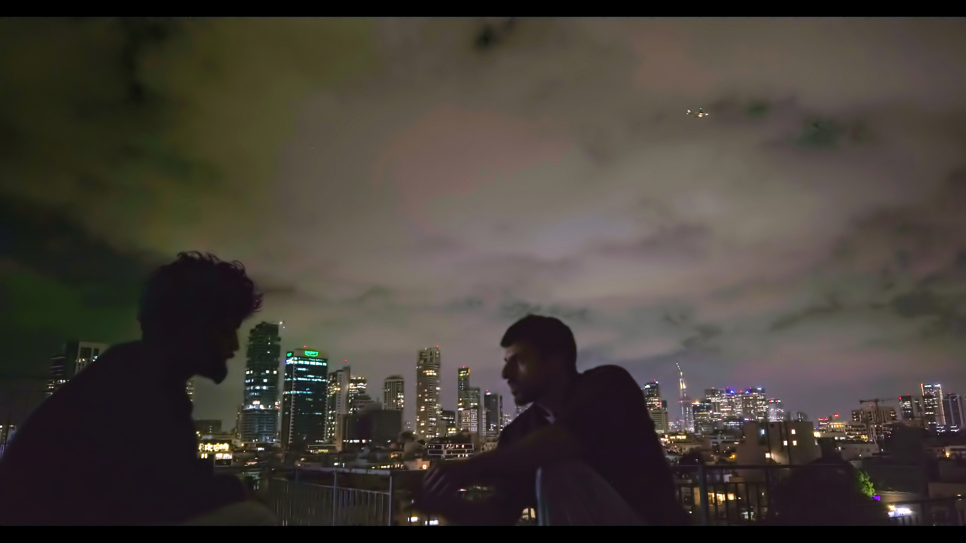Zwei Männer sitzen abends vor der Skyline von Tel Aviv mit Hochhäusern. Sie sind nur als dunkle Silhouetten zu sehen.