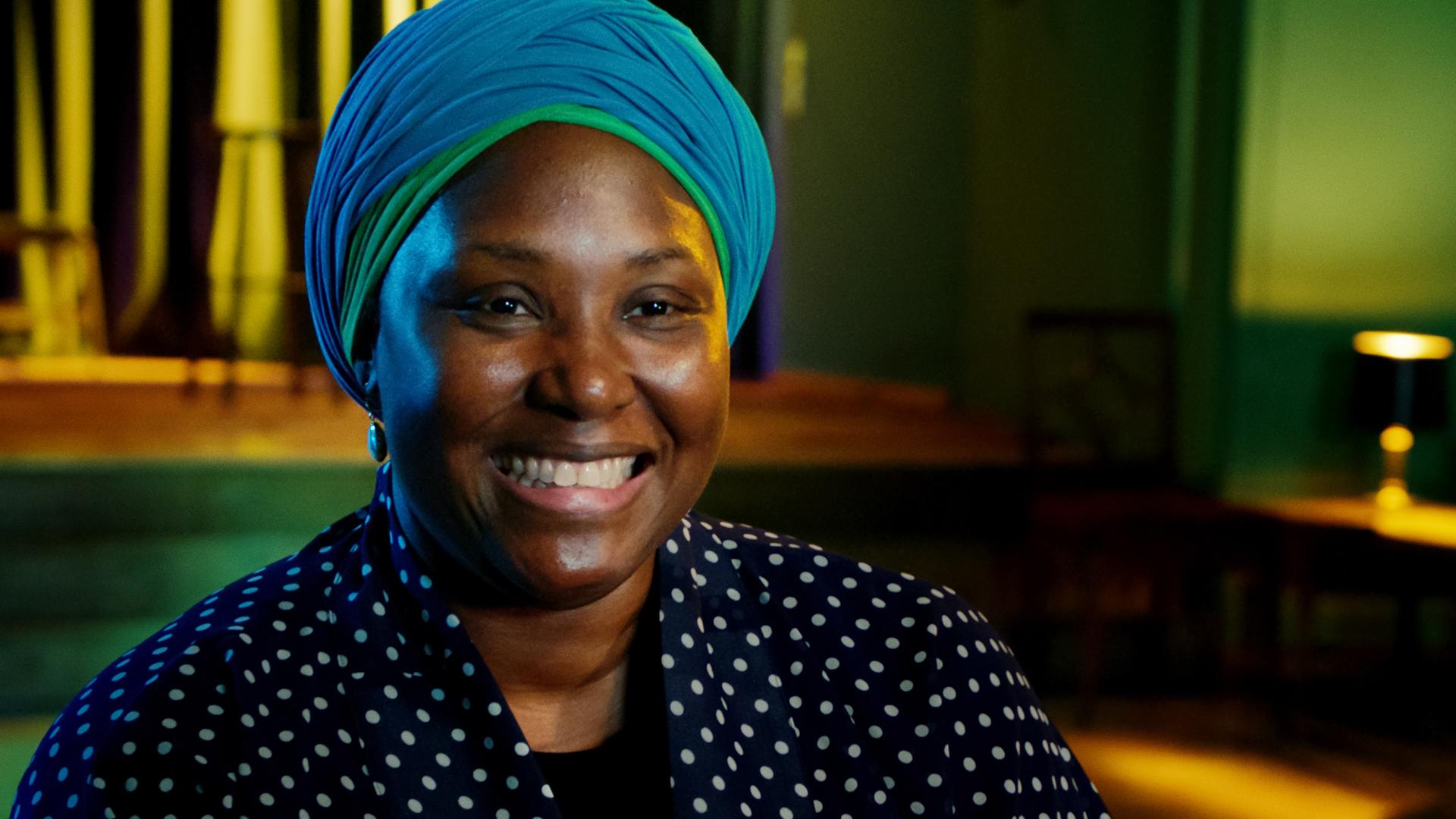 Man sieht eine schwarze Frau, die eine gepunktete Bluse trägt und einen blauen Turban auf dem Kopf.