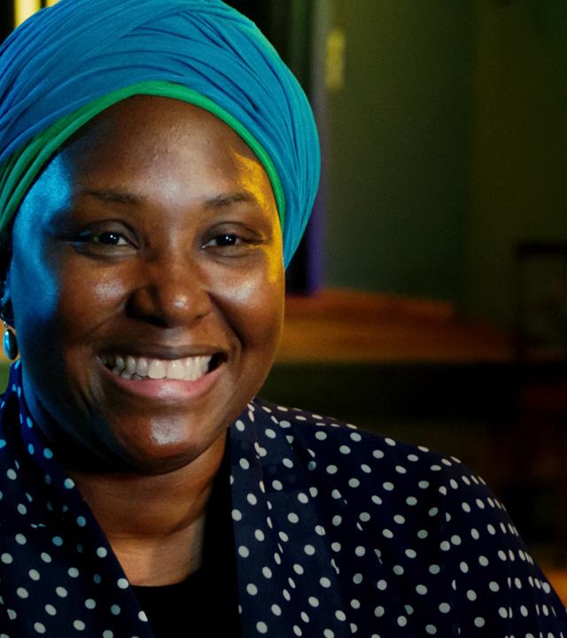 Man sieht eine schwarze Frau, die eine gepunktete Bluse trägt und einen blauen Turban auf dem Kopf.