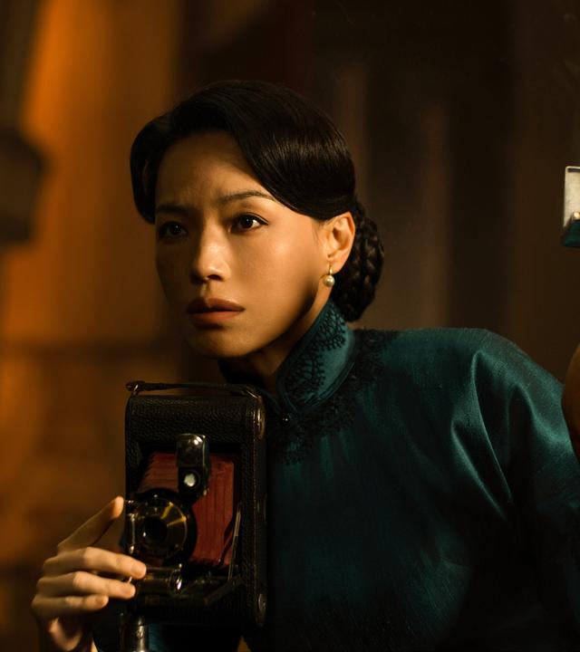 Eine dunkelhaarige Frau bedient einen historischen Fotoapparat, während sie ein Blitzlicht in einer Hand hoch hält.