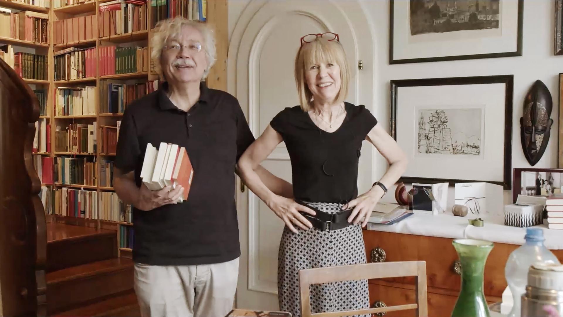Ein Mann und eine Frau stehen in einem Raum mit Bücherregal im Hintergrund.