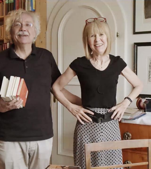 Ein Mann und eine Frau stehen in einem Raum mit Bücherregal im Hintergrund.