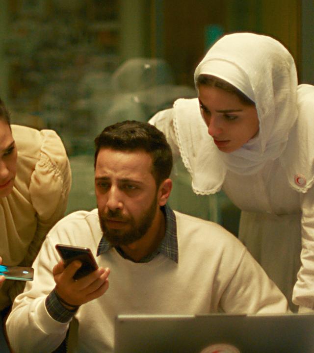 Ein Mann sitzt an einem Schreibtisch vor einem Laptop und er hält ein Handy in der Hand, in das er spricht. Sein Blick ist traurig und verzweifelt. Hinter ihm stehen drei Frauen, die sich besorgt vorbeugen und zuhören.