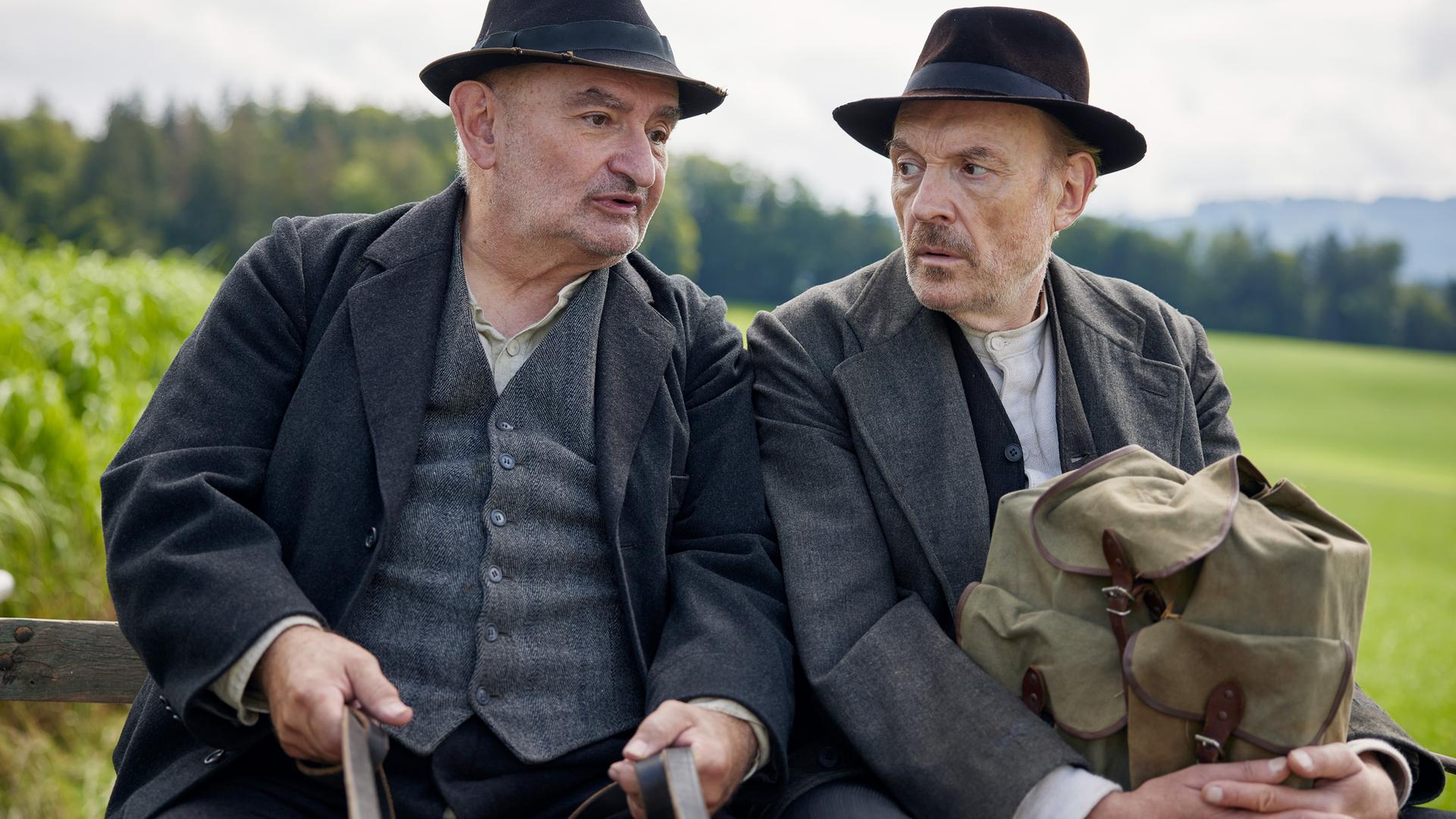 Zwei Männer in Anzügen und Hüten im Stil der 1920er Jahre sitzen nebeneinander auf einem Pferdewagen. Einer hält die Zügel, der andere einen Rucksack aus grobem Stoff.