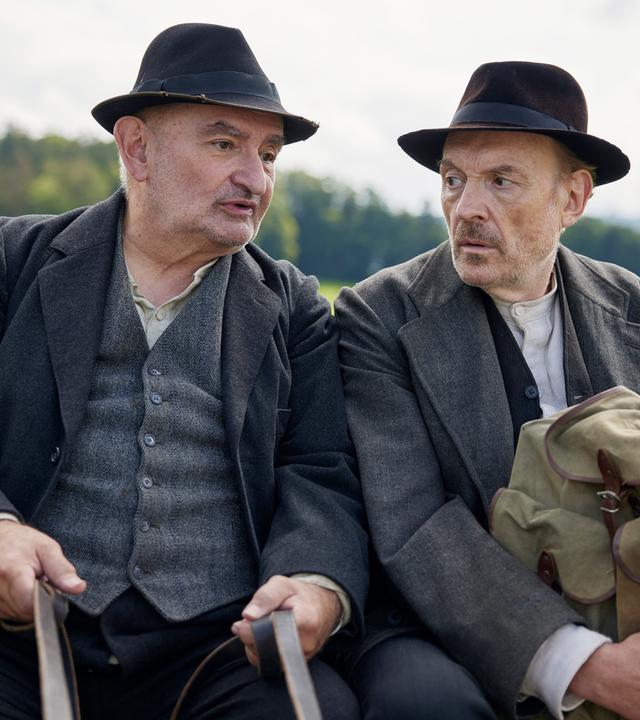 Zwei Männer in Anzügen und Hüten im Stil der 1920er Jahre sitzen nebeneinander auf einem Pferdewagen. Einer hält die Zügel, der andere einen Rucksack aus grobem Stoff.