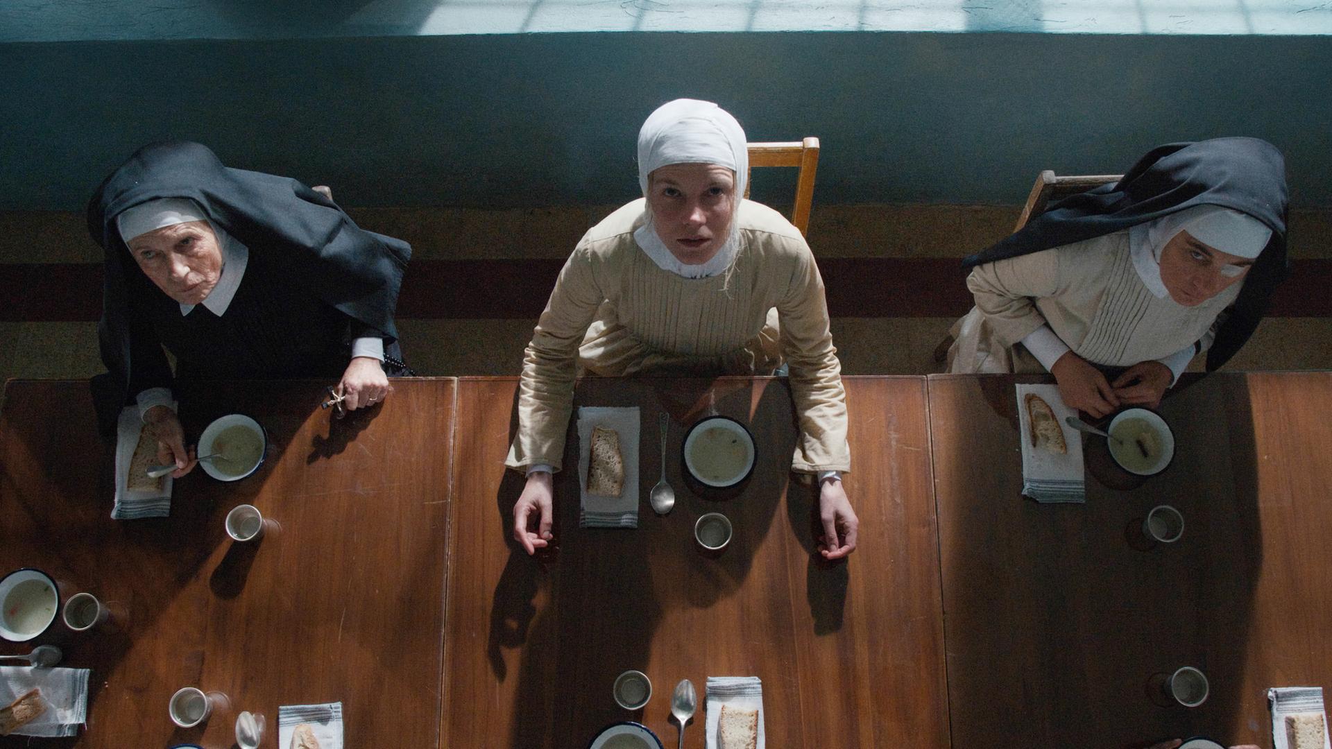 Blick von schräg oben auf drei Nonnen, die an einem Tisch sitzen und Teller vor sich haben.
