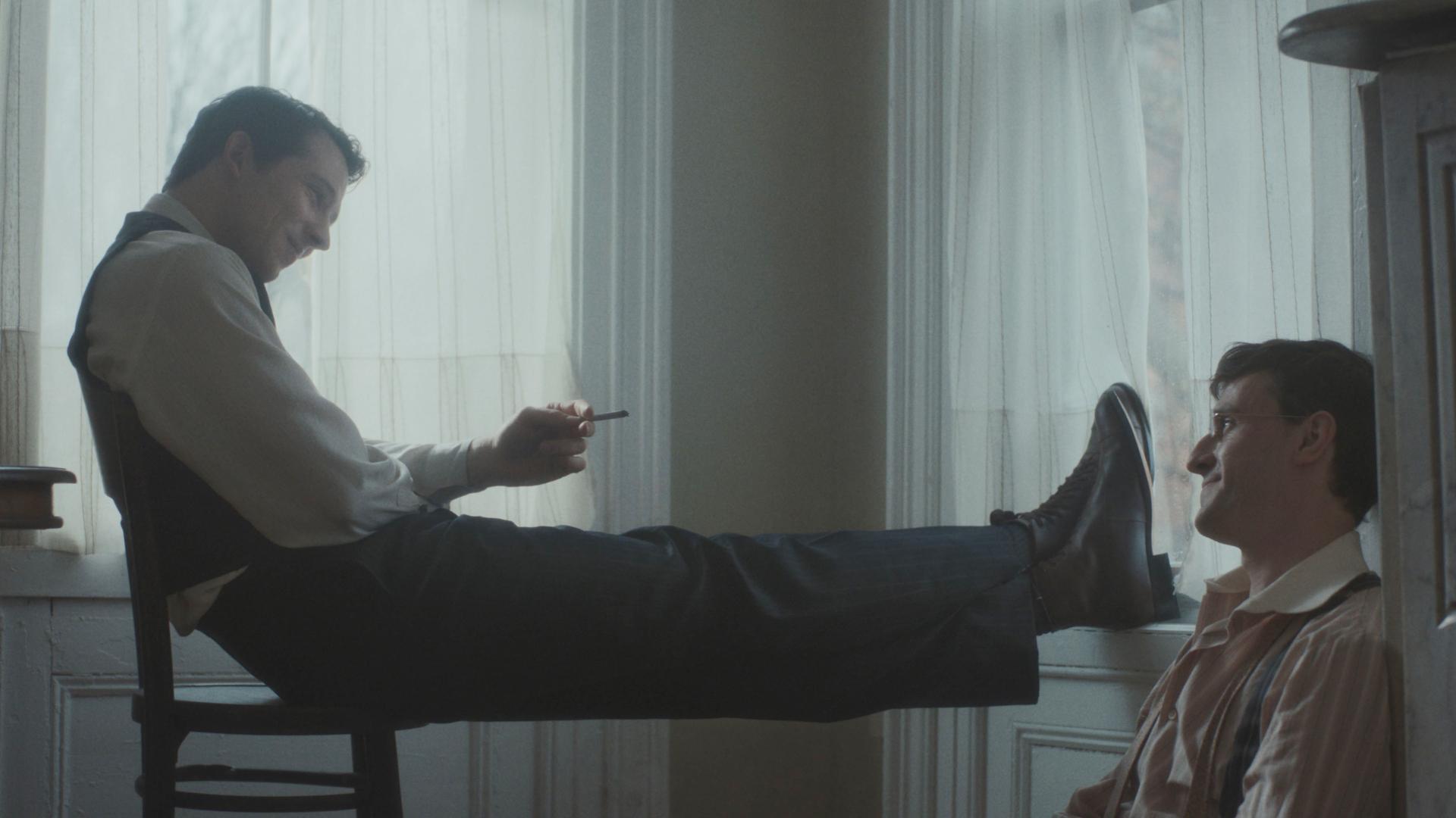 Ein junger Mann sitzt auf einem Stuhl, die Beine hochgelegt und eine Zigarette in der Hand, und schaut einen anderen jungen Mann an, der auf dem Boden an die Wand gelehnt sitzt. Die Männer lächeln einander an.