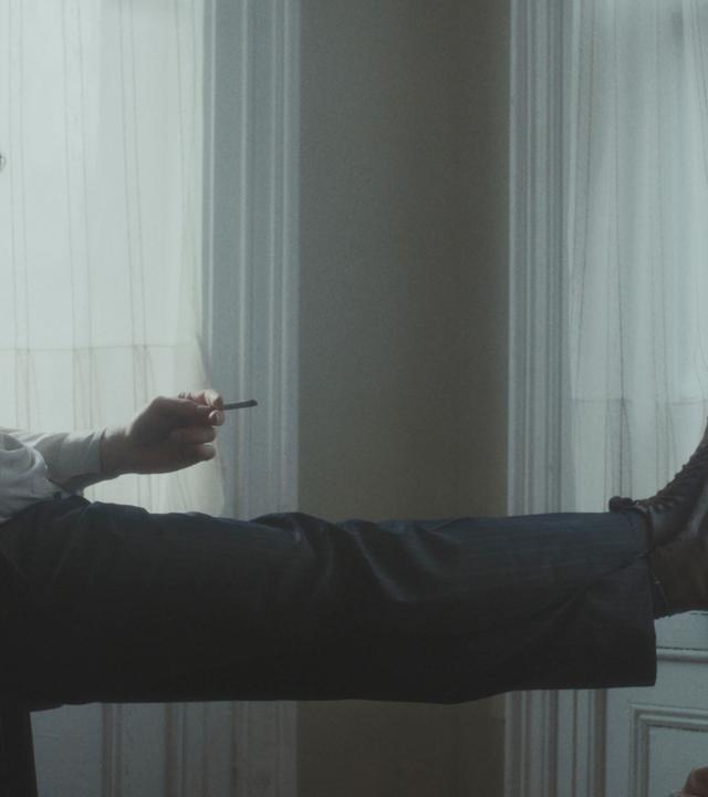 Ein junger Mann sitzt auf einem Stuhl, die Beine hochgelegt und eine Zigarette in der Hand, und schaut einen anderen jungen Mann an, der auf dem Boden an die Wand gelehnt sitzt. Die Männer lächeln einander an.