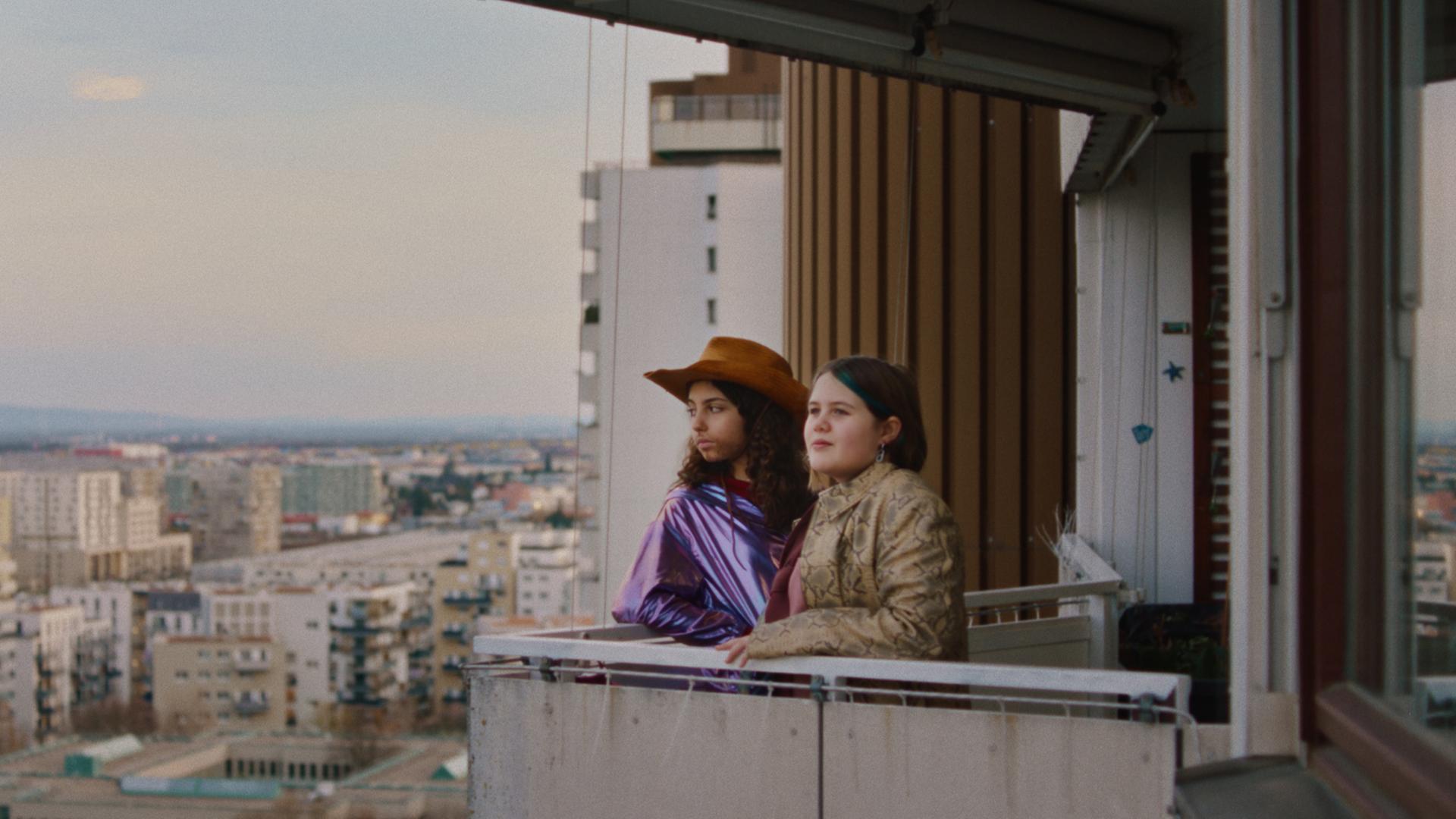 Zwei Mädchen stehen auf dem Balkon eines Hochhauses. Eine trägt einen Cowboyhut.