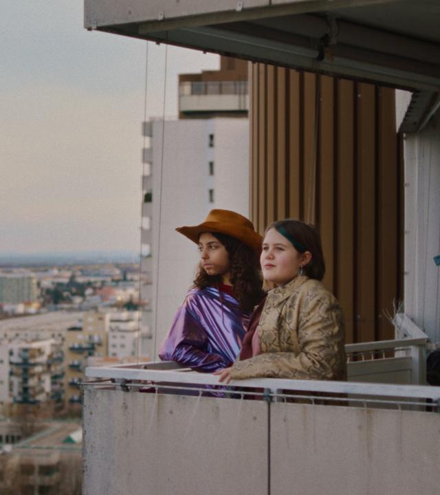 Zwei Mädchen stehen auf dem Balkon eines Hochhauses. Eine trägt einen Cowboyhut.