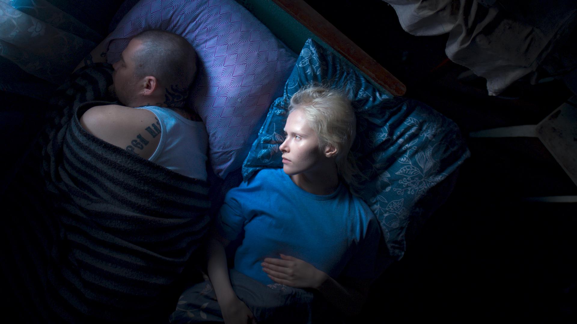 Eine junge Frau mit kurzen weißen Haaren liegt im Bett und betrachtet einen Mann mit kurzgeschorenen Haaren und Tattoos, der von ihr abgewendet neben ihr liegt und schläft.