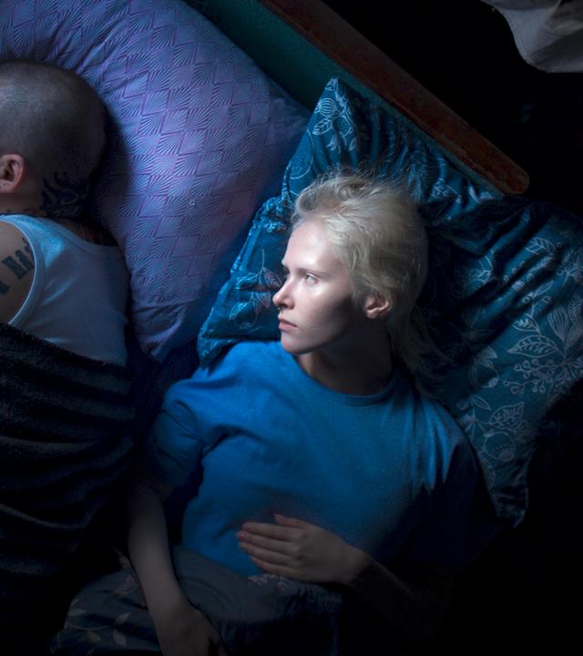 Eine junge Frau mit kurzen weißen Haaren liegt im Bett und betrachtet einen Mann mit kurzgeschorenen Haaren und Tattoos, der von ihr abgewendet neben ihr liegt und schläft.