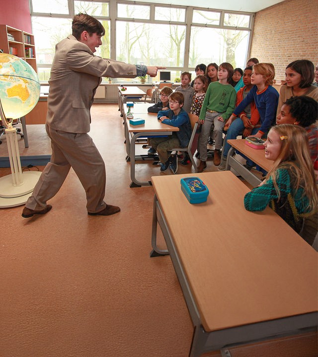 "Mister Twister" (Willem Voogd, li.) steht links im Klassenraum vor den gespannt auf ihn schauenden Schülern (rechts).