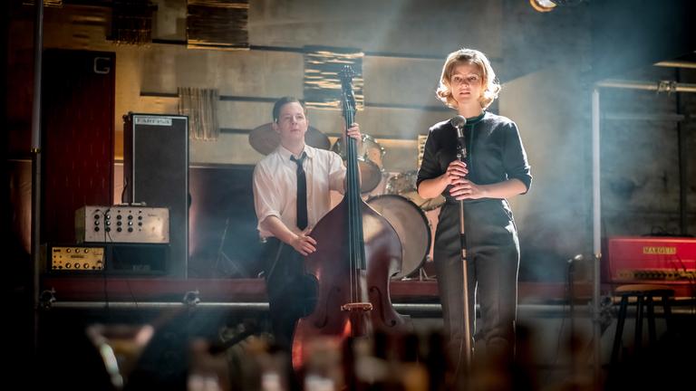 Monika (Sonja Gerhardt, r.) und Freddy (Trystan Pütter, l.) stehen auf einer Bühne. Monika hält ein Mikro in ihren Händen, während Freddy mit einem Cello neben ihr steht.