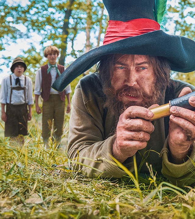 Die Jungen Kasperl und Seppel (Hintergrund) haben sich von hinten an den Räuber Hotzenplotz herangeschlichen und sehen erstaunt aus, ihn gefunden zu haben. Der Räuber, mit dreckiger Kleidung und großem Schlapphut, liegt im Vordergrund auf dem Boden und betrachtet das Messer in seiner Hand.