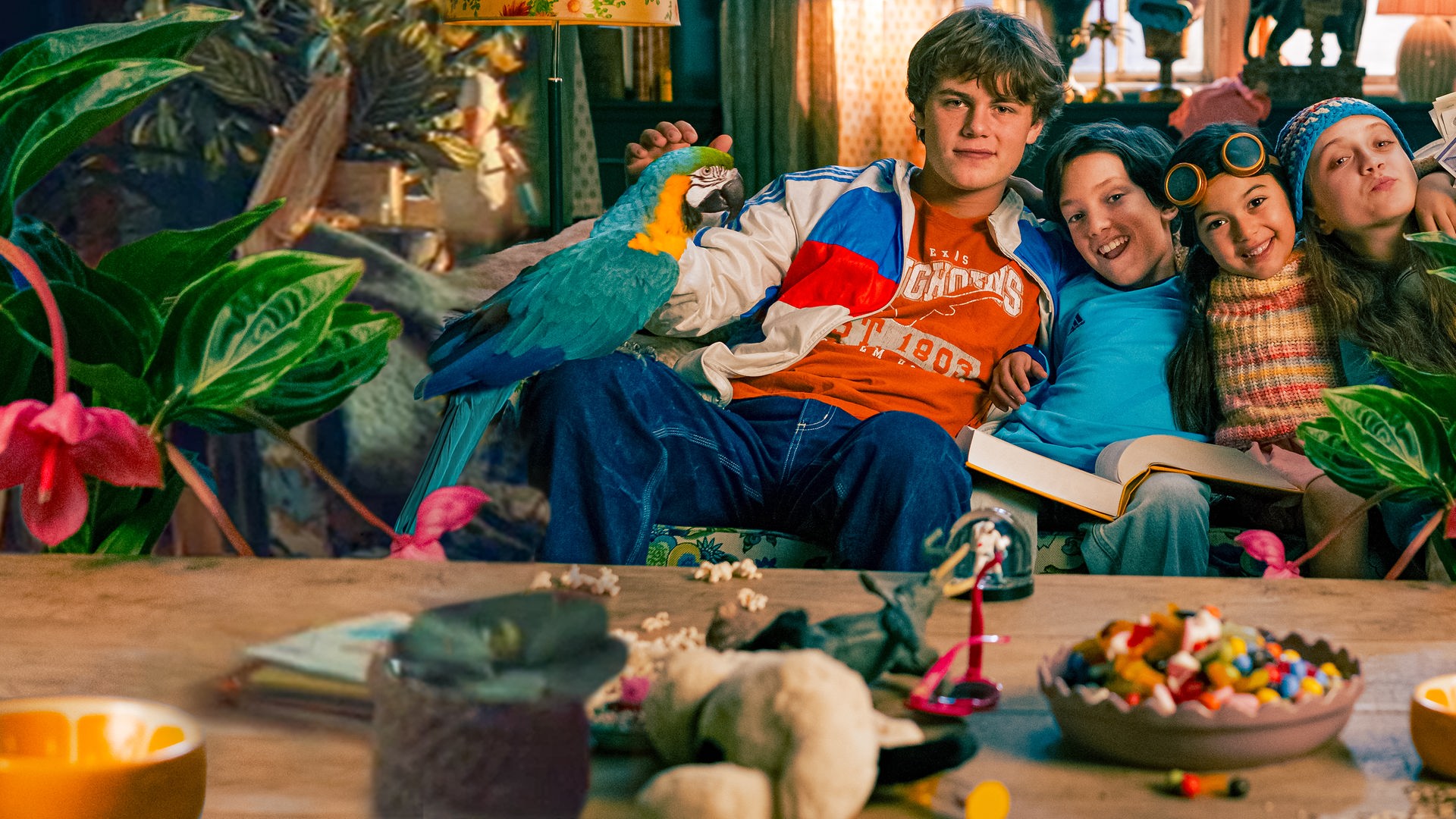 Wohnung: Tristan, Kirk, Petra und Katinka sitzen zusammen mit ihrem Papagei eng beieinander auf ihrem Sofa und umarmen sich. Sie sind bunt gekleidet und schauen fröhlich in die Kamera. Tristan streichelt den Papagei, Kirks dickes Lieblingsbuch liegt auf seinem Schoß, Petra trägt ein Fliegerbrille auf der Stirn und Katinka hält Geldscheine in die Luft.