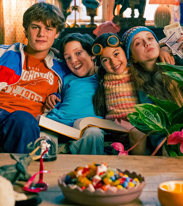 Wohnung: Tristan, Kirk, Petra und Katinka sitzen zusammen mit ihrem Papagei eng beieinander auf ihrem Sofa und umarmen sich. Sie sind bunt gekleidet und schauen fröhlich in die Kamera. Tristan streichelt den Papagei, Kirks dickes Lieblingsbuch liegt auf seinem Schoß, Petra trägt ein Fliegerbrille auf der Stirn und Katinka hält Geldscheine in die Luft.